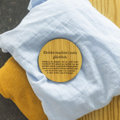 Holzmünze mit Text "Kleider machen Leute (glücklich).", welche auf Kleidung liegt.