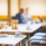 Eine Tasse und ein Teller mit Besteck stehen auf einer weißen Tischplatte. Im Hintergrund sieht man verschwommen weitere Tische und Personen.