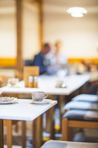 Eine Tasse und ein Teller mit Besteck stehen auf einer weißen Tischplatte. Im Hintergrund sieht man verschwommen weitere Tische und Personen.