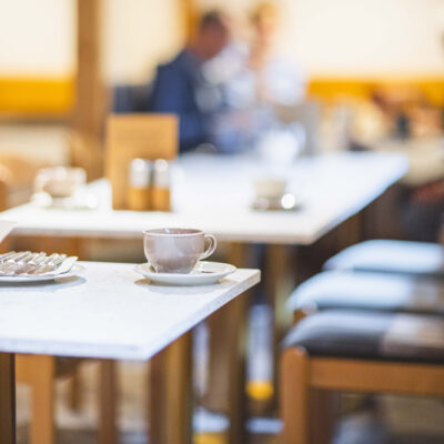 Eine Tasse und ein Teller mit Besteck stehen auf einer weißen Tischplatte. Im Hintergrund sieht man verschwommen weitere Tische und Personen.